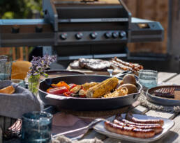 Outdoor barbecue scene with a large grill in the background and a cast-iron pan of corn, peppers, and vegetables on a wooden table.