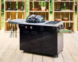 Black outdoor gas grill with hood on a wooden deck, adjacent prep area and hanging utensils on the side.