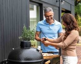 Couple smoking a grill outside, woman seasoning food while man watches and smiles nearby.