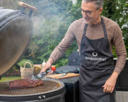Man grilling a slab of meat on a round barbecue grill outdoors, using a meat thermometer while wearing a black apron that says 'outdoorchef'.